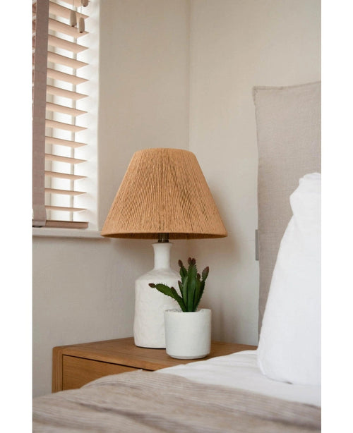 Junto a una cama con ropa de cama blanca y beige, junto a una ventana con persianas de madera, se encuentra una mesita de noche con una lámpara blanca con pantalla tejida de color beige y una planta suculenta artificial de 20 cm.