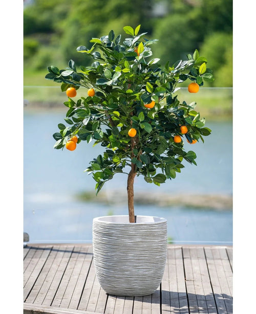 Un naranjo artificial de 120 cm con frutos maduros se encuentra en una maceta con relieve blanco en una terraza de madera, con vistas a una fuente de agua brumosa y un paisaje verde en el exterior.