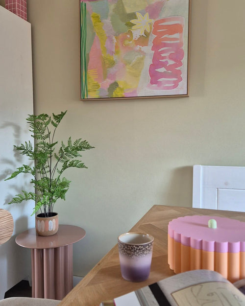 En una sala de temática pastel hay una planta de helecho artificial de 85 cm sobre una mesa auxiliar rosa, una taza morada y dorada, un cuenco rosa con el borde festoneado, un cuaderno abierto y en la pared un cuadro abstracto con tonos verdes, rosas y amarillos.