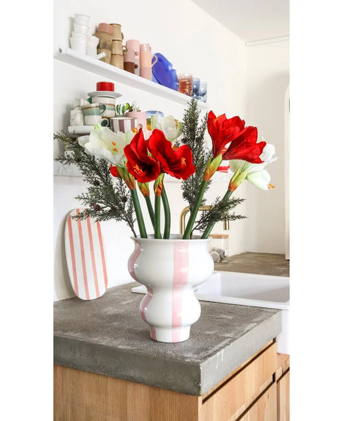 La flor artificial Amaryllis Real Touch roja de 36 cm se encuentra en un jarrón de cerámica blanco con rayas rosas sobre una encimera de hormigón en una cocina luminosa con estantes y vajilla de colores al fondo.