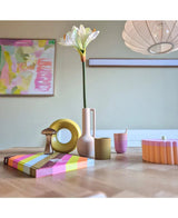 Una moderna mesa de madera muestra objetos coloridos, incluido un jarrón rosa que contiene la flor artificial Amaryllis Real Touch blanca de 66 cm, una escultura de un anillo amarillo, libros a rayas y una taza rosa. El arte abstracto acentúa la pared del fondo.