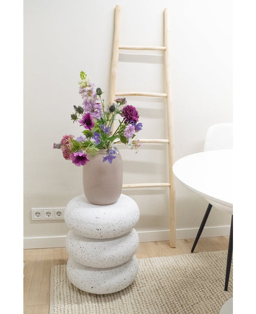 Una flor artificial de campanilla de 62 cm de color violeta se encuentra en un jarrón sobre tres cojines redondos que parecen piedras blancas. Detrás hay una escalera de madera clara contra una pared blanca, junto a una mesa y una silla blancas sobre una alfombra beige.