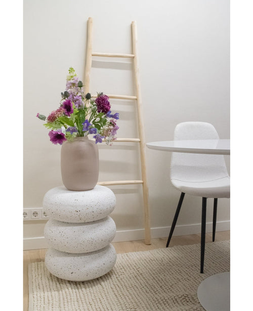 Una flor artificial de campanilla violeta de 62 cm se encuentra en un jarrón beige sobre taburetes de terrazo blanco apilados. Detrás hay una escalera de madera clara, cerca de una silla blanca y una mesa redonda, todo ello colocado sobre una alfombra beige texturizada en una habitación minimalista.