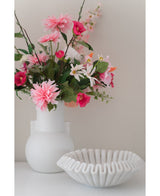 Una flor artificial Dalia de 68 cm de color rosa en un jarrón blanco se encuentra junto a un cuenco de cerámica blanca con volantes sobre una superficie blanca, sobre un fondo liso y claro.