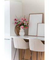 Una moderna cocina blanca con dos taburetes de bar beige, una flor artificial Dalia rosa de 68 cm en un jarrón blanco sobre la encimera y dos obras de arte abstractas enmarcadas contra la pared.
