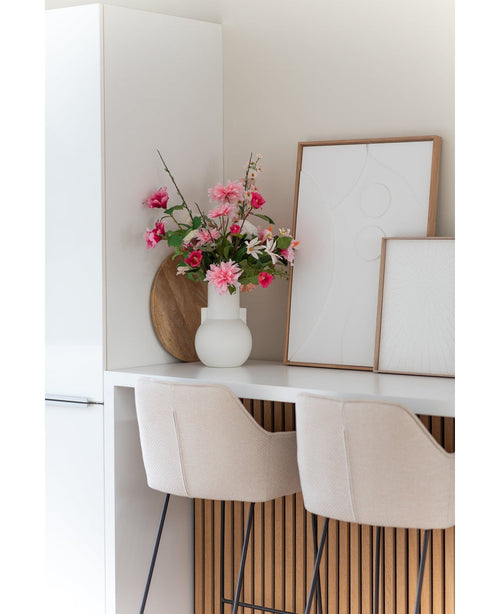 Una moderna cocina blanca con dos taburetes de bar beige, una flor artificial Dalia rosa de 68 cm en un jarrón blanco sobre la encimera y dos obras de arte abstractas enmarcadas contra la pared.