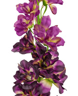 Un primer plano de la Flor Artificial Delphinium Púrpura de 79 cm, un tallo artificial alto con múltiples flores de color violeta y violeta oscuro con un corazón verde y amarillo, sobre un fondo blanco.