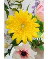 La flor artificial Gerbera de 53 cm amarilla, una flor artificial de color amarillo vivo con pétalos en capas, destaca entre otras coloridas flores artificiales y hojas verdes, con flores blancas y rosas de fondo.