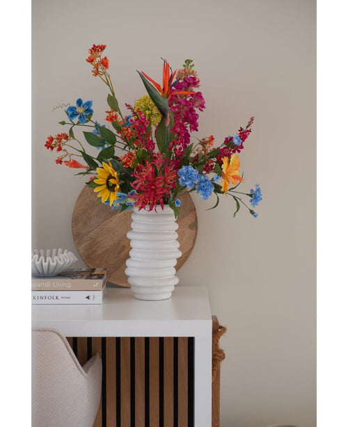 Un jarrón blanco texturizado sostiene un ramo de flores artificiales de hierba gatera rosa de 75 cm sobre una mesa blanca junto a libros apilados y decoración, con una bandeja redonda de madera apoyada contra la pared beige al fondo.