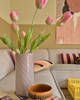 Una flor artificial de tulipán francés de 63 cm en color rosa se encuentra en un jarrón sobre la mesa junto a una taza naranja en una acogedora sala de estar con un sofá color crema, cojines estampados, una caja amarilla y arte abstracto en la pared al fondo.