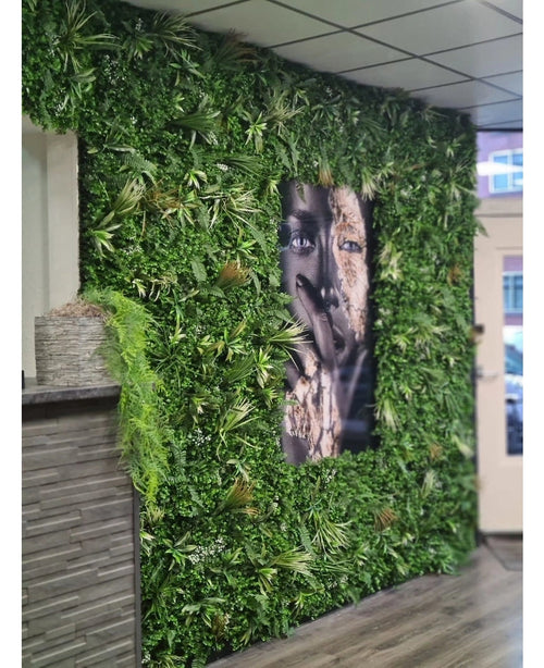 Un panel ignífugo Kunsthaag Savanne de 100x100 cm con exuberante vegetación rodea un gran retrato facial en blanco y negro en un espacio interior con paneles de techo de azulejos y un pequeño mostrador a la izquierda.