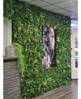 Una pared cubierta con un seto artificial Savanne de 100x100 cm, con vegetación protegida contra los rayos UV en una maceta de plástico de Ø100, tiene en el centro un gran retrato en blanco y negro. El interior moderno tiene un techo de tejas y un mostrador de madera a la izquierda.