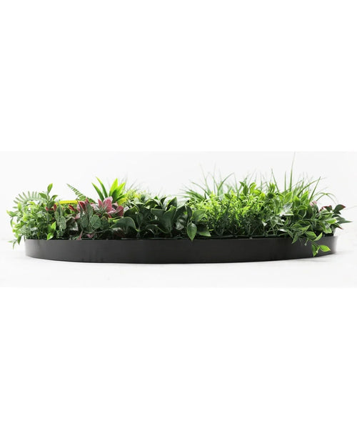 El seto artificial con marco negro Ø100 cm es una jardinera semicircular negra llena de exuberante follaje verde y algunas hojas teñidas de rosa, sobre un fondo blanco.