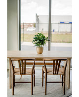 Una mesa de madera con dos sillas a juego se encuentra sobre una alfombra redonda cerca de grandes ventanales. Una Planta Artificial Baby Schefflera UV 45 cm decora la mesa, con vistas a los edificios y a una valla exterior.
