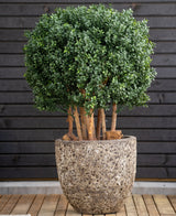 La bola de boj artificial de madera estilo romano UV 90 cm, una planta redonda de hoja verde con tallos gruesos, se encuentra en una gran maceta de piedra sobre madera, contra una pared horizontal oscura con paneles de madera.