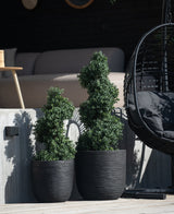 Dos plantas topiarias artificiales en espiral de boj estilo romano UV de 120 cm en maceteros con textura negra se encuentran afuera sobre un escalón de concreto, con una mecedora moderna y un sofá beige visibles al fondo.