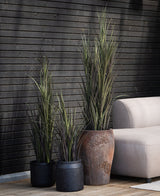 Tres plantas artificiales de Césped Artificial Burdeos UV de 90 cm con largas y esbeltas hojas verdes se encuentran junto a un sofá beige sobre una terraza de madera, contra una pared de listones de madera oscura.