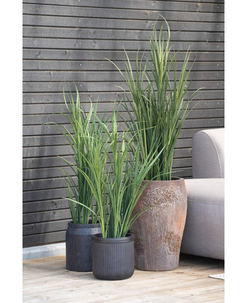 Tres plantas artificiales de césped artificial verde UV de 150 cm con hojas largas y estrechas se encuentran sobre una terraza de madera junto a un sofá de exterior gris claro, contra una pared de listones de madera oscura.