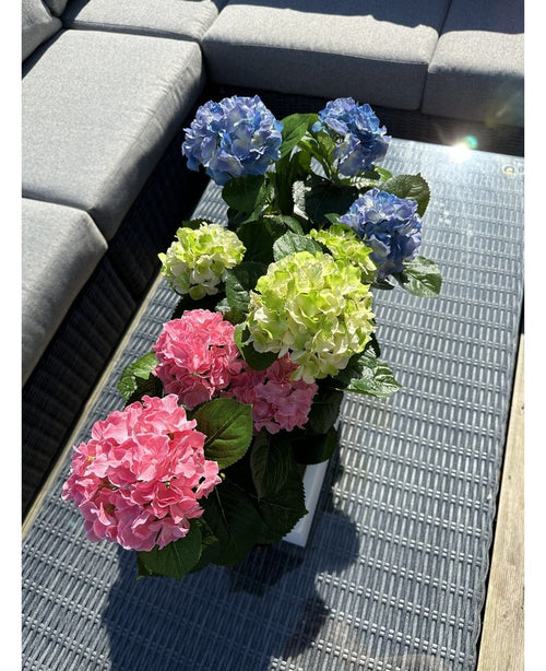 La planta Hortensia Artificial Verde UV 35 cm se encuentra sobre una mesa de mimbre con tapa de cristal junto a un sofá de exterior gris, con hortensias verdes realistas que se iluminan con la luz del sol.