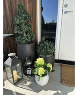 En la terraza, cerca de una puerta marrón, hay una planta artificial de hortensia verde UV de 35 cm en una maceta blanca con flores de color amarillo verdoso, junto a dos plantas en una maceta negra y dos faroles decorativos con velas.
