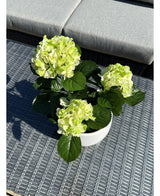 La hortensia artificial verde UV de 35 cm, con flores de color verde claro y hojas de color verde oscuro, se encuentra sobre una mesa de exterior tejida de color gris con un banco acolchado al fondo.