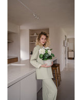 Una mujer de pelo rubio rizado, vestida con un traje verde claro, se encuentra en una cocina moderna con una planta Artificial Hydrangea White UV 35 cm en sus manos – con flores de color blanco crema – y sonríe a la cámara.