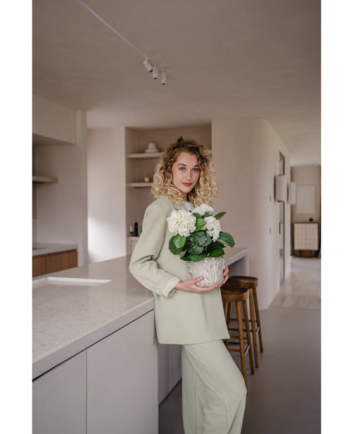 Una mujer de pelo rubio rizado, vestida con un traje verde claro, se encuentra en una cocina moderna con una planta Artificial Hydrangea White UV 35 cm en sus manos – con flores de color blanco crema – y sonríe a la cámara.