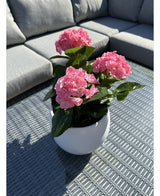 La Planta Artificial Hortensia Rosa UV 35 cm, una maceta blanca con flores de hortensias de color rosa vibrante, se alza sobre una mesa de exterior tejida en gris con un sofá gris con cojines al fondo, iluminada por la luz del sol y sombras suaves.