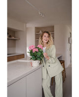 Una mujer con un traje verde claro se encuentra en una cocina moderna y sonríe mientras sostiene la planta Artificial Hydrangea Pink UV de 35 cm sobre el mostrador.