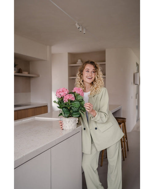 Una mujer con un traje verde claro se encuentra en una cocina moderna y sonríe mientras sostiene la planta Artificial Hydrangea Pink UV de 35 cm sobre el mostrador.
