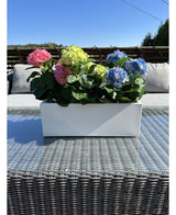 La Planta Artificial Hortensia Rosa UV 35 cm, una jardinera rectangular blanca con hortensias rosas, se coloca en el exterior sobre una mesa de mimbre con tapa de cristal, cojines grises y un cielo azul de fondo.