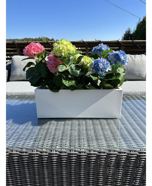 La Planta Artificial Hortensia Rosa UV 35 cm, una jardinera rectangular blanca con hortensias rosas, se coloca en el exterior sobre una mesa de mimbre con tapa de cristal, cojines grises y un cielo azul de fondo.