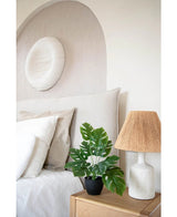 Un dormitorio moderno en tonos neutros tiene una cama blanca con almohadas beige y blancas, una mesita de noche de madera con una lámpara de cerámica y una pantalla tejida, arte abstracto en la pared encima de la cama y Art Monstera de 40 cm en una maceta decorativa negra que agrega vegetación.