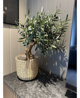 El soporte de olivo artificial UV de 60 cm, con hojas verdes y aceitunas negras, se coloca en una cesta tejida sobre una superficie de mármol oscuro contra una pared con paneles claros: fácil de mantener y resistente a los rayos UV para una belleza duradera.
