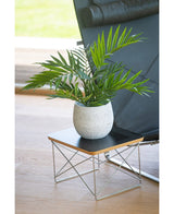 Una palmera artificial UV de 60 cm se encuentra en una maceta sobre una pequeña mesa auxiliar moderna con tablero negro y patas de alambre metálico, junto a una silla negra sobre un suelo de madera clara.