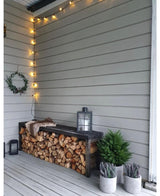 Un acogedor rincón al aire libre tiene un banco de leña apilada, una linterna encima, la planta artificial Conifer deluxe UV 60 cm, plantas en macetas cerca, una corona en la pared y luces de cadena encima, contra las paredes de madera gris claro.