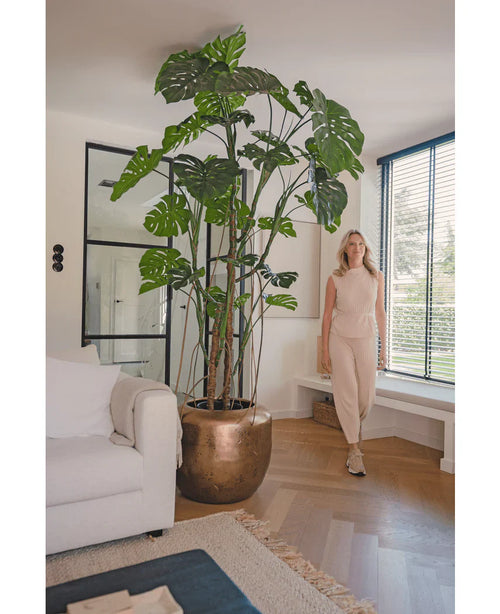 Una mujer vestida de beige está junto a la planta Monstera artificial de 240 cm en una maceta de bronce. El moderno salón tiene un sofá blanco, grandes ventanales con persianas y suelo de madera.