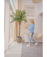 Una mujer con blusa azul y vaqueros pasa por el Kunstpalm Phoenix De Luxe 250 cm en un salón luminoso y moderno con ventanas altas, decoración neutra y obras de arte enmarcadas en la pared.