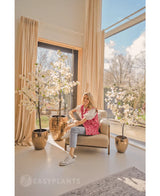 Una mujer se ríe en un sillón beige cerca de grandes ventanales con cortinas beige, rodeada por tres buganvillas artificiales de 150 cm blancas en un espacio luminoso, moderno y lleno de luz natural.