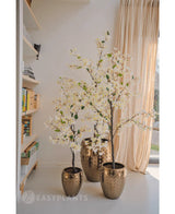Una planta de buganvilla artificial que mide 150 cm de altura se encuentra en una maceta dorada decorativa cerca de una ventana iluminada por el sol con cortinas beige y agrega elegancia al espacio acogedor. Las estanterías cercanas realzan el acogedor ambiente interior.