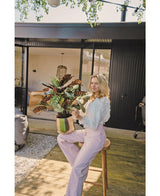 Una mujer rubia con una blusa blanca y pantalones de color violeta claro se sienta afuera en un taburete con la planta en maceta roja artificial Calathea Makoyana UV de 75 cm, en una terraza de madera frente a una casa moderna con puertas de vidrio.
