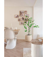 Un comedor moderno y minimalista con sillas blancas, una mesa de madera y la planta artificial Enkianthus de 150 cm que añade vegetación, complementado con una galería de fotografías enmarcadas sobre un fondo de color neutro.
