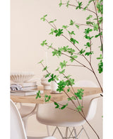 Una moderna zona de comedor con sillas blancas y una mesa de madera clara, decorada con libros, velas y un jarrón blanco, está enmarcada en primer plano por la planta artificial de hoja verde Enkianthus de 150 cm.