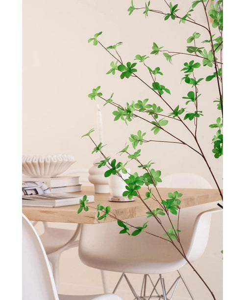 Una moderna zona de comedor con sillas blancas y una mesa de madera clara, decorada con libros, velas y un jarrón blanco, está enmarcada en primer plano por la planta artificial de hoja verde Enkianthus de 150 cm.