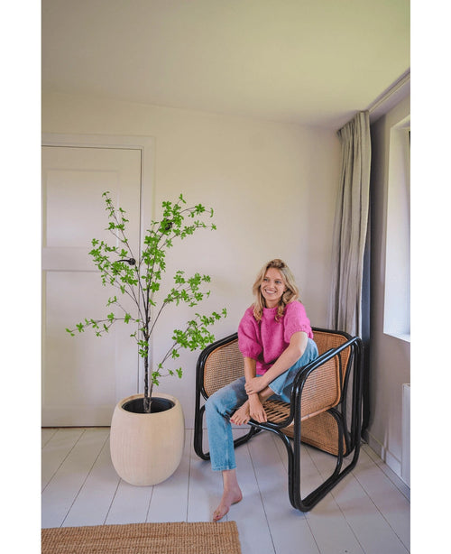 Una mujer con un top rosa y vaqueros azules está sentada descalza en una moderna silla de mimbre junto a la planta artificial Enkianthus de 150 cm en una habitación luminosa y minimalista con paredes blancas y alfombras claras.