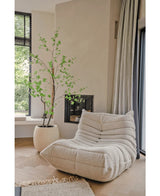 Un acogedor sillón texturizado de color crema se encuentra junto a la planta artificial Enkianthus de 210 cm y una moderna chimenea en una sala de estar luminosa y minimalista con suelos de madera clara y grandes ventanales.