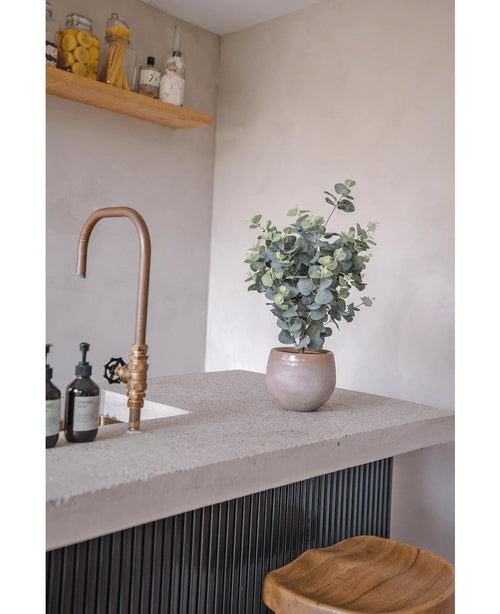 Una encimera moderna con grifo de bronce, botes de jabón, un taburete de madera y el Eucalipto Artificial de 76 cm. Al fondo, sobre una pared gris claro, se encuentran estantes de madera con tarros y botellas.