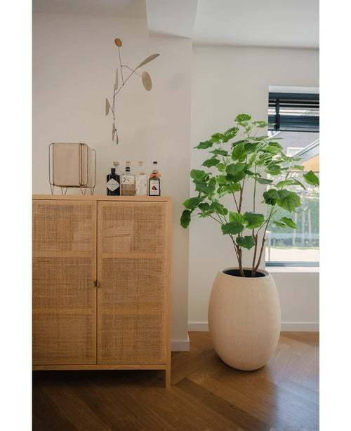 Un armario de madera clara con botellas y decoración se encuentra junto a una planta Ficus artificial de 150 cm en una maceta de color crema cerca de una ventana con persianas, en una habitación moderna y luminosa con suelo de madera.