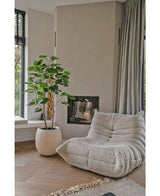 Un acogedor sillón beige con estructura se encuentra junto a la planta artificial Ficus de 150 cm en una sala de estar moderna con suelos de madera clara, paredes neutras, una ventana y cortinas grises hasta el suelo.