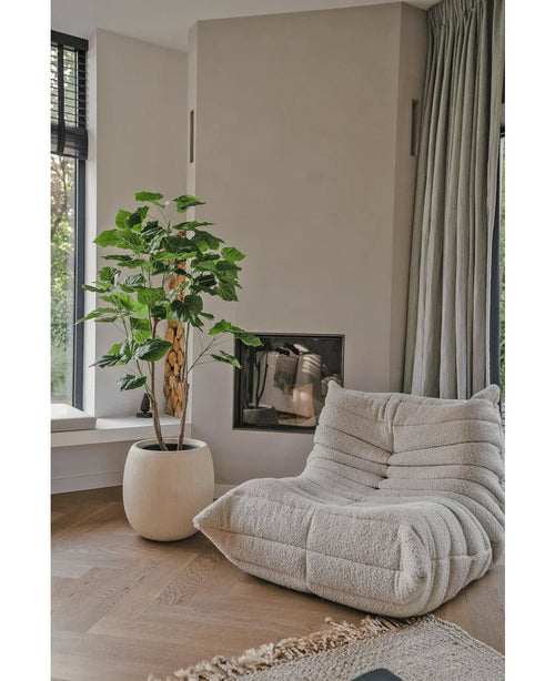 Un acogedor sillón beige con estructura se encuentra junto a la planta artificial Ficus de 150 cm en una sala de estar moderna con suelos de madera clara, paredes neutras, una ventana y cortinas grises hasta el suelo.